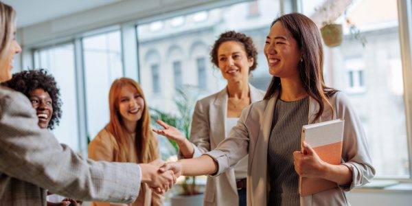 New asian intern holding a notebook while handshaking women from her team at work in the lounge